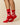 A person wearing Warm & Wonderful Sheep Socks Diana Red—red crew socks with white sheep and one black sheep—stands on tiptoe against a plain beige background.