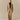 A woman stands in a studio wearing Rowing Blazers Cath Herringbone Pant Camel, paired with an oversized jacket, black cropped top, and black open-toe shoes against a plain beige background.