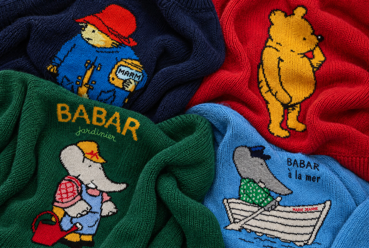 Four knitted baby socks with colorful cartoon characters, including Paddington Bear, Winnie the Pooh, Babar the elephant, and a dolphin in a boat, arranged in a flat-lay pattern.