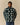 A person wears a forest green Warm & Wonderful Sheep Sherpa Jacket Green, featuring an all-over pattern of small white sheep with one solitary black sheep on the chest.