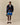 A man holds a green cap, wearing the Rowing Blazers x Paddington™ Paddington Sherpa Jacket Navy, a light blue button-down shirt, blue shorts, white socks, and brown loafers against a beige background.