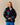 A woman with blond hair wears a Rowing Blazers x Paddington™ Paddington Sherpa Jacket Navy with colorful cartoon graphics, a white shirt, and blue shorts against a plain beige background.