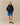 A person stands in a studio wearing the Rowing Blazers x Paddington™ Paddington Sherpa Jacket Navy, paired with blue shorts and black shoes. The background is plain beige.