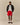 A person wearing a Tuxedo Paddington Classic Sherpa Black jacket over a white graphic t-shirt, red athletic shorts, white crew socks, and brown leather boat shoes.