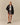 A person wearing a Rowing Blazers x Paddington™ Tuxedo Paddington Classic Sherpa Black jacket, a white graphic t-shirt, and a plaid pleated skirt.