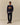 A man wears the Rowing Blazers Cotton NY Hot Dog Sweater Navy over a white collared shirt, paired with dark jeans and brown shoes, standing against a plain beige background.