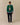 A man wears a green Rowing Blazers x Babar Babar Jardinier Cashmere Sweater with a Babar the Elephant intarsia motif over a light blue button-down shirt and navy trousers.