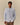 A man wears the Paddington Oxford Light Blue Stripe shirt by Rowing Blazers x Paddington™ over a white top with brown pants, standing against a plain beige background.