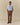 A man faces forward, wearing the Paddington Oxford Light Blue Stripe shirt by Rowing Blazers x Paddington™ over a white graphic tee, brown pleated trousers, and dark loafers, hands in pockets against a plain beige background.