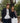 A man and woman posing against a stone railing. The man is wearing a navy colored blazer with a white, grey, and cream colored sweater underneath and blue pants. The woman is wearing a blue and red plaid blazer with a navy sweater underneath and white pants. There are trees in the background.