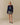 A model wears a navy Rowing Blazers x Paddington™ Penelope Paddington Cashmere Sweater featuring an intarsia Paddington Bear design, paired with a pink collared shirt and a black and white patterned pleated skirt.