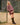 A blonde woman wearing a deep red sweater with a white sheep pattern. It is layered on top of a maroon plaid dress with a fringe. She is wearing black knee high boots and leaning against a fence.