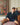 A man sitting on a bed leaning against the wall. He is wearing a green and navy argyle sweater. There is a colorful painting on the wall.